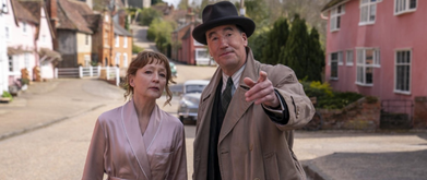 a detective in a hat and trench coat pointing to something down the street next to a woman in a pink silk bathrobe. 